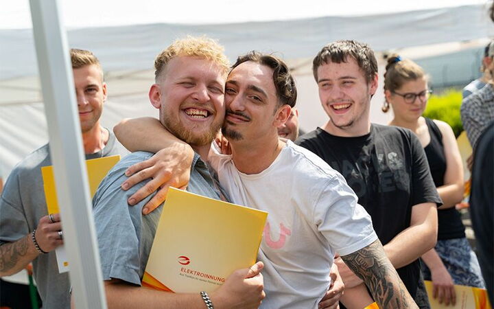 Mehrere junge Menschen, darunter zwei Männer, umarmen sich lachend und halten Mappen der Elektroinnung.
